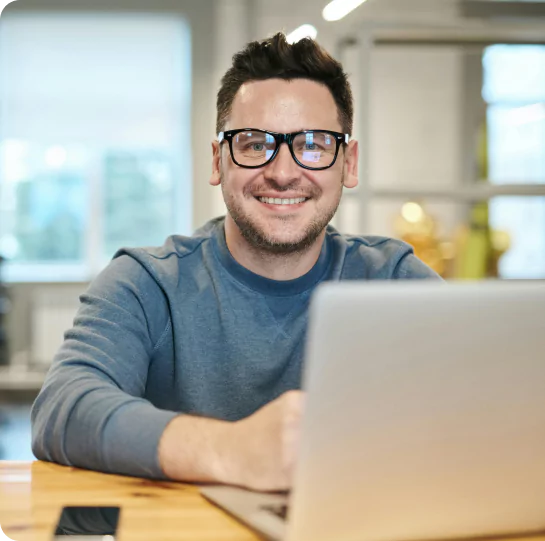 Retrato de um empreendedor sorridente com óculos, sentado à secretária em frente a um computador portátil, representando a satisfação do cliente com os serviços de contabilidade e apoio à gestão da Mundiempresarial.